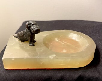 May include: A small, rectangular ashtray made of translucent, light-yellow material with a rounded indentation. A small, dark-colored dog figurine sits on the left side. The ashtray has a reddish-brown base.