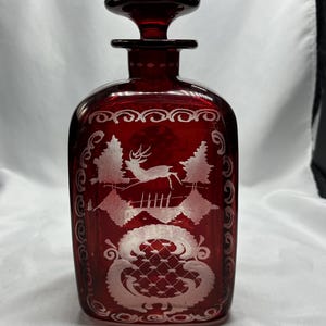May include: A ruby red glass decanter with a stopper. The decanter features an etched design of a deer leaping between trees and decorative scrollwork. The stopper is a matching red color.