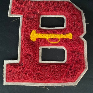 May include: A red felt letter B with a white outline and a yellow trumpet design in the center. The letter is a vintage style patch.
