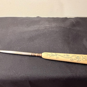 May include: A button hook with a long, slender metal shaft ending in a curved hook. The handle is made of a smooth, cream-colored material featuring a delicate, dark-inked line drawing of floral vines and leaves etched onto its surface.