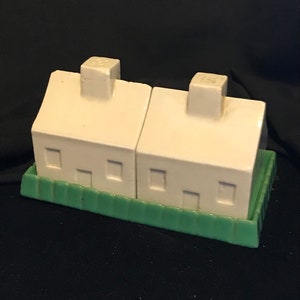 May include: A set of two white ceramic salt and pepper shakers shaped like houses with green trim. The shakers are sitting on a green base.
