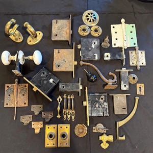 May include: A collection of antique hardware components, including door knobs, hinges, latches, and hooks. The items are in various metallic finishes, such as brass, bronze, and black, with signs of age and wear. The assortment suggests a vintage or restoration theme.