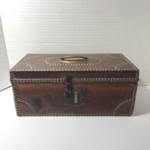 May include: A rectangular brown leather storage box featuring a brass handle on the lid and a central metal latch mechanism. The exterior is decorated with rows of small brass studs along the edges and corners, creating a structured appearance.