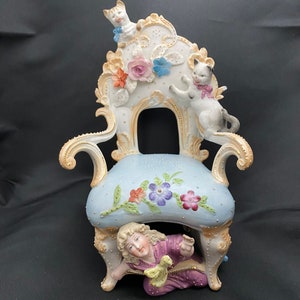 May include: A porcelain figurine of a girl hiding under a chair with two cats on top. The chair is decorated with flowers and gold accents. The girl is wearing a purple dress and has blonde hair.
