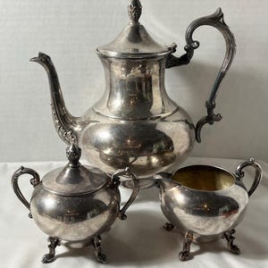 May include: A silver-plated tea service set consisting of a large teapot with a lid, a lidded sugar bowl, and an open creamer. Each piece features a polished metallic finish, ornate curved handles, and decorative feet on the smaller vessels.