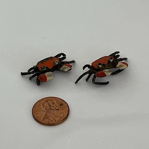 May include: Two small, detailed crab figurines featuring dark brown bodies with vibrant orange shells and white-tipped claws. A copper-colored United States one-cent coin is positioned below the crabs to provide a clear sense of their small scale.