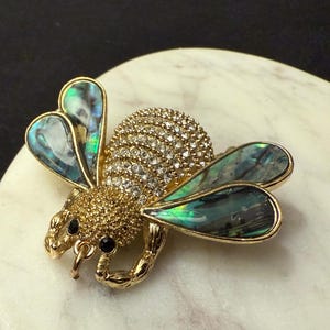 May include: A gold-toned bee brooch with a body encrusted with clear crystals. The wings are iridescent, displaying shades of green, blue, and gold. The brooch is set on a white and gray marble surface.