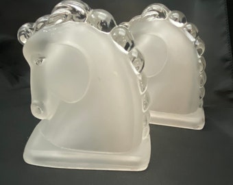 May include: A pair of frosted glass bookends shaped like horse heads. The bookends have a textured surface and are a light white color.