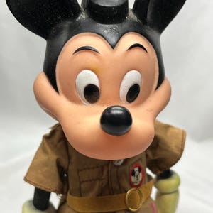 May include: Vintage Mickey Mouse figurine wearing a brown uniform with a yellow belt and a circular buckle. The figurine has black ears, a black nose, and a tan face. The uniform has a patch with Mickey Mouse's image.