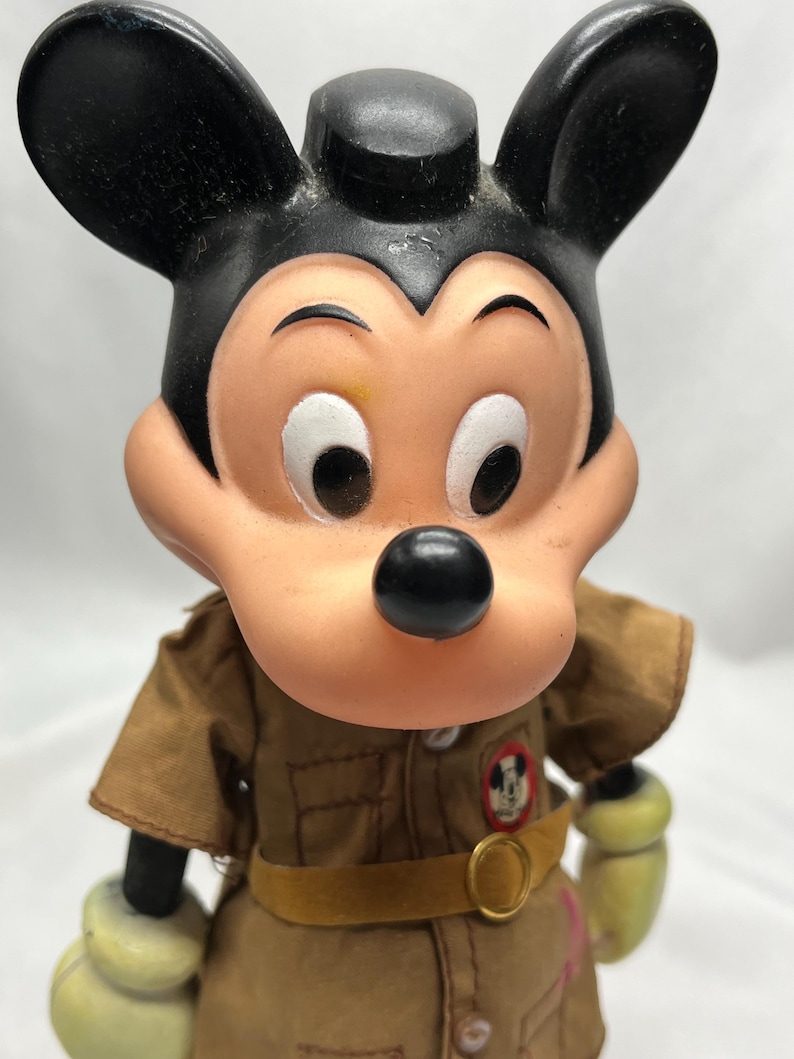 May include: Vintage Mickey Mouse figurine wearing a brown uniform with a yellow belt and a circular buckle. The figurine has black ears, a black nose, and a tan face. The uniform has a patch with Mickey Mouse's image.