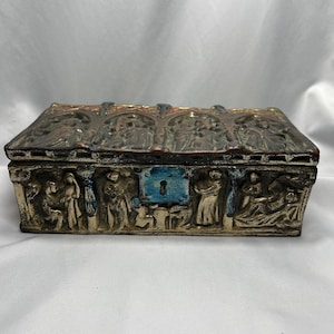 May include: A rectangular antique box with a hinged lid. The box is made of metal and has a silver finish. The lid is decorated with a relief design of figures and arches. The box has a blue keyhole.
