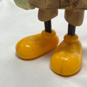 May include: Close-up of a doll's legs and feet. The doll has bright yellow shoes and tan-colored legs. The doll is wearing a brown coat. The background is a white surface.