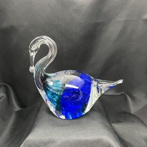 May include: A clear glass swan figurine with a long, curved neck and a blue and teal body. The glass has a swirling pattern of colors and is displayed on a black surface.