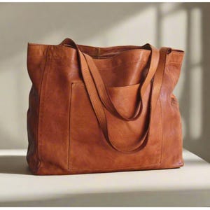Peut inclure: Un grand sac cabas rectangulaire en cuir lisse de couleur cognac. Le sac est doté de deux longues anses robustes et d'une grande poche extérieure profonde centrée sur le panneau avant, présentant une finition naturelle et mate.