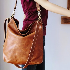 May include: A large, slouchy brown leather hobo bag featuring a spacious main compartment, a wide shoulder strap, and antique brass-toned metal hardware including swivel clasps and rivets. The bag has a smooth, matte finish and a structured base.