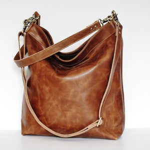May include: A medium brown leather hobo bag with a smooth, slightly distressed finish. The bag features a single main compartment, a top zipper closure, and an adjustable shoulder strap attached with antique brass-toned metal swivel clasps.