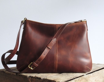 Può includere: Una borsa a tracolla in pelle color marrone rossiccio di medie dimensioni con finitura liscia, una cucitura verticale centrale e una tracolla regolabile con fibbia in metallo color ottone. La borsa presenta un'apertura superiore curva e cuciture rinforzate.