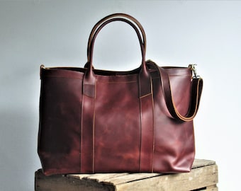 May include: A deep burgundy leather tote bag featuring two sturdy top handles and a detachable shoulder strap. The bag has a rectangular silhouette with visible stitching along the vertical panels and metallic hardware clips on the sides.