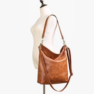 May include: A medium brown hobo-style shoulder bag crafted from a smooth, textured material. The bag features a slouchy silhouette, a single main compartment, and a long, adjustable strap attached with metallic hardware clips on both sides.