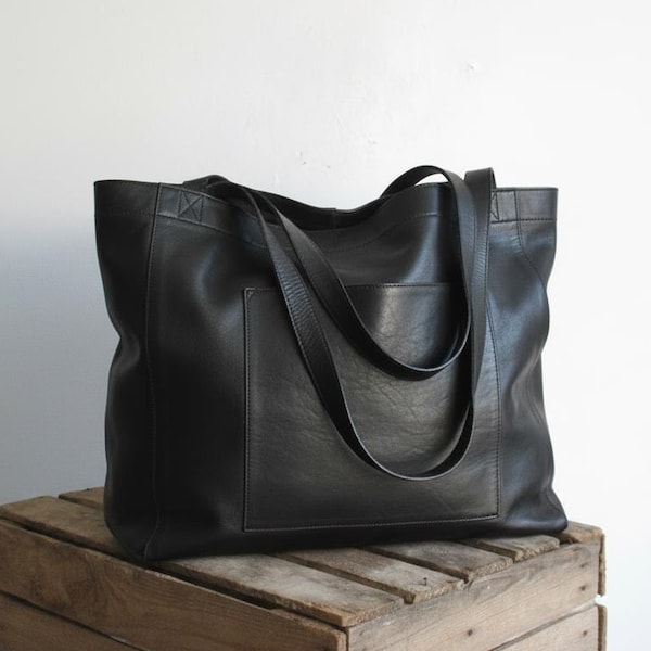 May include: A black leather tote bag featuring a large rectangular front pocket, two long shoulder straps, and visible stitching along the top edges and pocket seams. The bag has a structured rectangular shape with a smooth, matte finish.