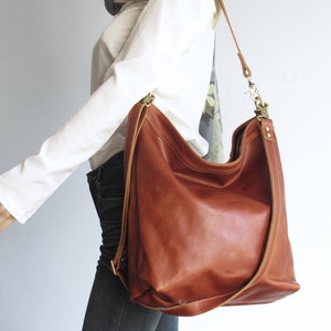 May include: A large, cognac-colored leather hobo bag featuring a slouchy, unstructured silhouette. The bag includes a long, adjustable shoulder strap attached with brass-toned metal hardware, visible stitching along the edges, and a wide opening.