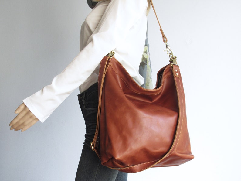 May include: A large, cognac-colored leather hobo bag featuring a slouchy, unstructured silhouette. The bag includes a long, adjustable shoulder strap attached with brass-toned metal hardware, visible stitching along the edges, and a wide opening.