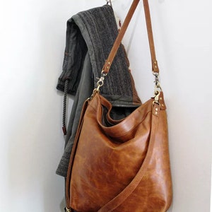 May include: A large, slouchy hobo-style shoulder bag crafted from smooth, cognac-brown leather. The bag features a long, adjustable shoulder strap attached with brass-toned metal swivel clips and rivets, providing a flexible, unstructured shape.