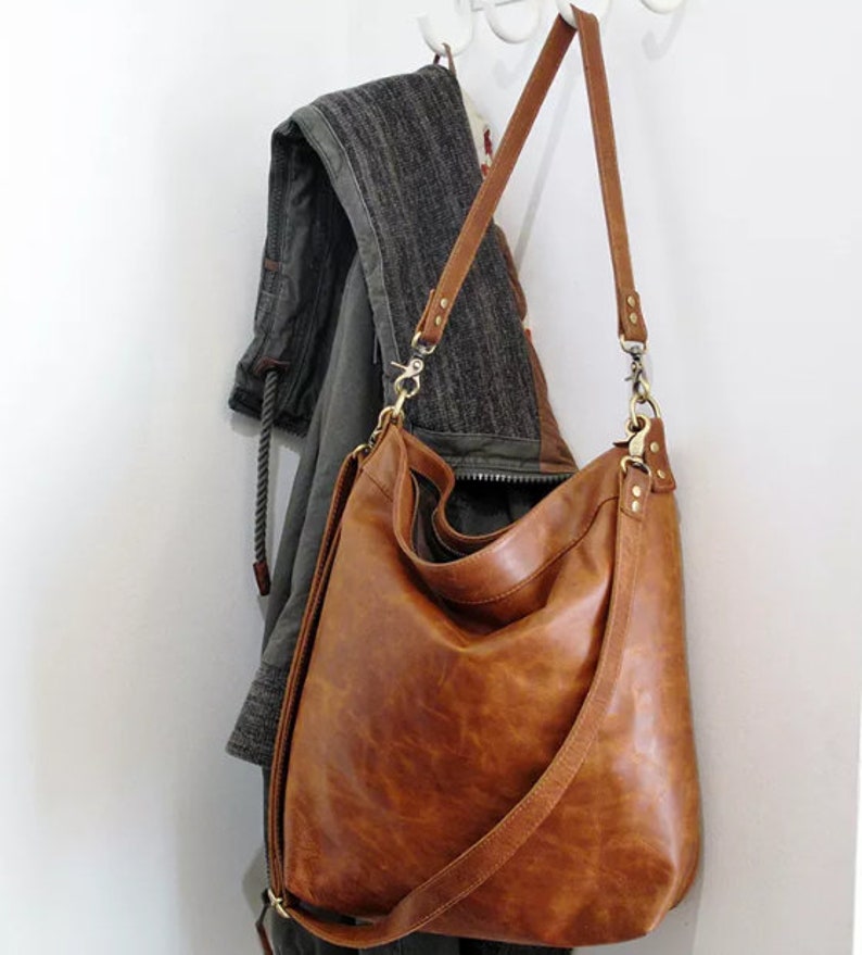 May include: A large, slouchy hobo-style shoulder bag crafted from smooth, cognac-brown leather. The bag features a long, adjustable shoulder strap attached with brass-toned metal swivel clips and rivets, providing a flexible, unstructured shape.