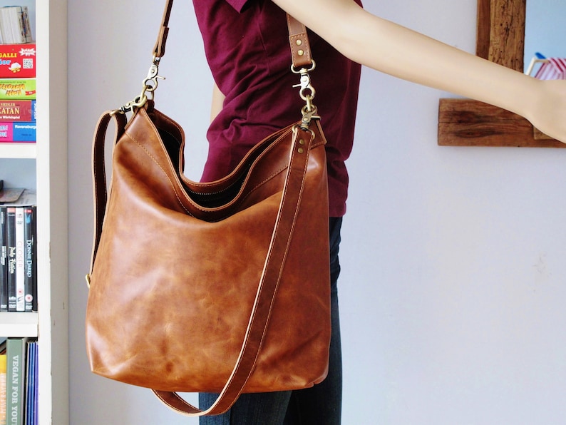 May include: A large, slouchy brown leather hobo bag featuring a spacious main compartment, a wide shoulder strap, and antique brass-toned metal hardware including swivel clasps and rivets. The bag has a smooth, matte finish and a structured base.