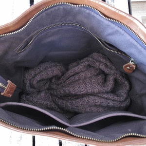 May include: A brown leather handbag with a dark gray fabric interior lining. The bag features a top zipper closure, an internal zippered pocket, and an open slip pocket. A dark gray knitted garment is folded and placed inside the main compartment.
