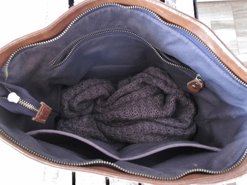 May include: A brown leather handbag with a dark gray fabric interior lining. The bag features a top zipper closure, an internal zippered pocket, and an open slip pocket. A dark gray knitted garment is folded and placed inside the main compartment.