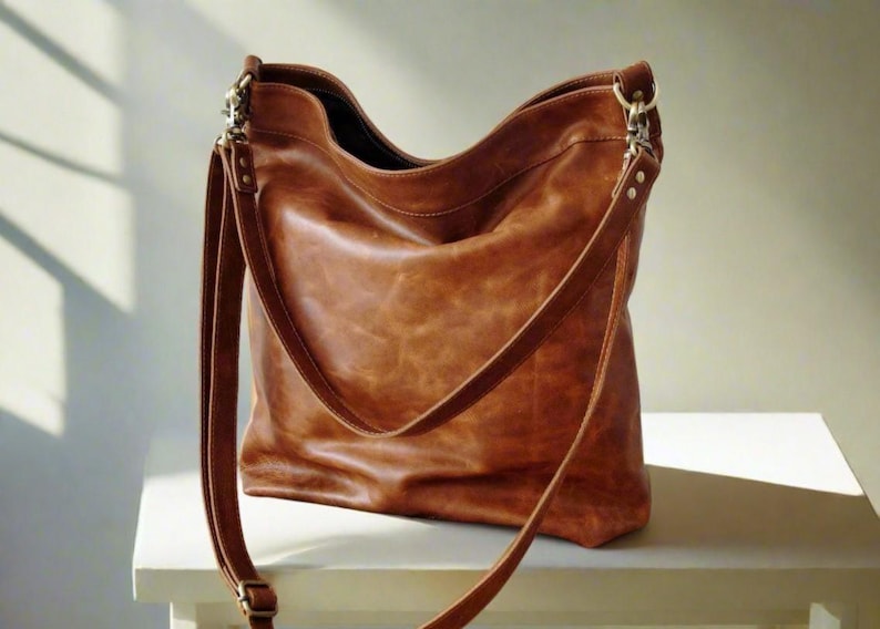 May include: A medium-brown leather hobo bag featuring a slouchy silhouette, a wide main compartment, and a long adjustable shoulder strap attached with silver-toned metal swivel clips and rivets. The surface displays a natural, textured grain.