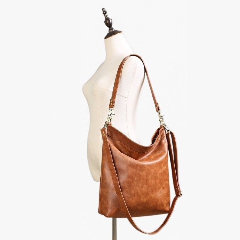 May include: A medium brown hobo-style shoulder bag crafted from a smooth, textured material. The bag features a slouchy silhouette, a single main compartment, and a long, adjustable strap attached with metallic hardware clips on both sides.