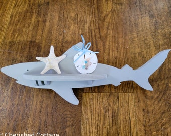 May include: A grey wooden shark shelf with a starfish and a sand dollar on the shelf. The shelf is mounted on a wood wall.
