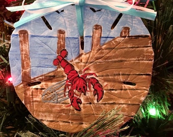 May include: A hand-painted sand dollar ornament featuring a red lobster on a wooden dock with blue water in the background. The ornament is tied with a blue and white ribbon.