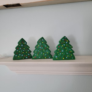 May include: Three green wooden Christmas trees with colorful painted ornaments. The trees are arranged in a row on a white shelf.