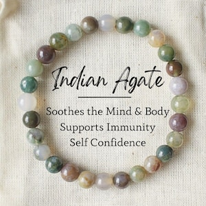 May include: A bracelet made of Indian agate beads in shades of green, brown, and white. The bracelet is on a white cloth bag with the text "Indian Agate" and "Soothes the Mind & Body, Supports Immunity, Self Confidence".