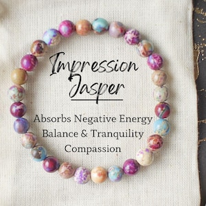 May include: A multi-colored Impression Jasper bracelet with a white background. The bracelet is made of small, round beads and has a tag that reads "Impression Jasper" and "Absorbs Negative Energy, Balance & Tranquility, Compassion."