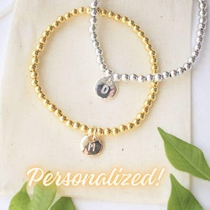 Könnte beinhalten: Zwei personalisierte Perlenarmbänder, eines golden und eines silbern, mit Initialanhängern. Die Armbänder liegen auf einem weißen Hintergrund mit grünen Blättern.