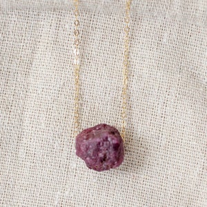 May include: A gold chain necklace with a rough, dark pink gemstone pendant. The necklace rests on a beige linen pouch. A card with a floral illustration and the text "the ROSEMARY room" is in the background.