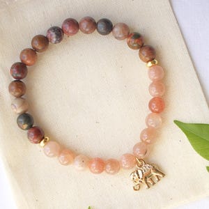 May include: A pink and brown gemstone bracelet with a gold elephant charm. The bracelet is on a white background with green leaves.