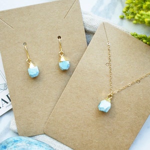 Puede incluir: Un conjunto de joyas delicadas con un collar de colgante de piedra azul claro y pendientes a juego. El collar y los pendientes se muestran en cartulina marr&oacute;n con el texto "the rosemar" visible.
