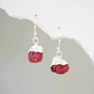 May include: A pair of silver earrings with a raw ruby gemstone dangle. The ruby is a deep red color and has a rough, natural texture.