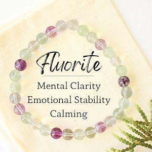 Puede incluir: Una pulsera de cuentas de Fluorita con cuentas verdes y moradas descansa sobre una bolsa de color crema. La imagen incluye el texto "Fluorita, Claridad Mental, Estabilidad Emocional, Calmante."
