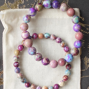 May include: Two bracelets made of colorful, multi-colored, and patterned beads. The bracelets are in a small white cloth bag. The bag is on a dark gray background with gold accents.