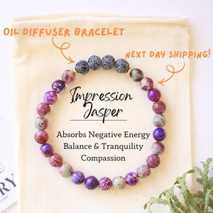 May include: An oil diffuser bracelet made of Impression Jasper beads in shades of purple, red, and green. The bracelet is displayed on a beige pouch with a card from The Rosemary Room. The text on the card reads "Absorbs Negative Energy, Balance & Tranquility, Compassion."