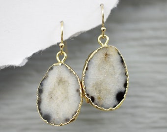 Puede incluir: Un par de pendientes de tono dorado con gotas de piedra de ágata blanca. Las piedras tienen un aspecto natural y orgánico con patrones en blanco y negro.