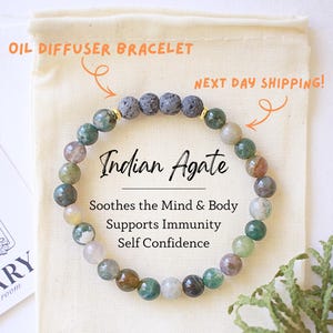 May include: An Indian Agate oil diffuser bracelet with green, gray, and brown beads. The bracelet is displayed on a cream-colored pouch with the text "Indian Agate" and "Soothes the Mind & Body, Supports Immunity, Self Confidence."