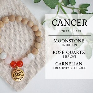 Puede incluir: Un brazalete con encanto del zodíaco de Cáncer con cuentas de madera, piedra lunar, cuarzo rosa y cuentas de cornalina. El brazalete está sobre un fondo blanco con una pequeña bolsa de tela y hojas verdes.