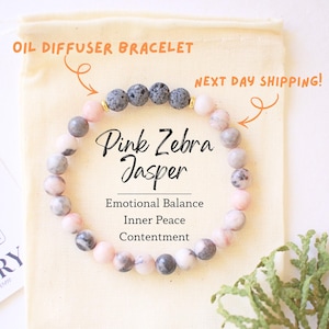 Op de afbeelding: Roze Zebra Jaspis olie diffuser armband met roze, grijze en zwarte kralen. De armband wordt getoond op een beige zakje met de tekst "Emotionele Balans, Innerlijke Vrede, Tevredenheid."